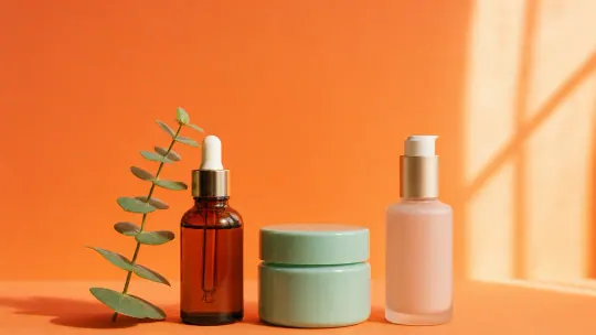 A young woman in a soft-lit bathroom holding up two different skincare bottles side by side and looking unsure